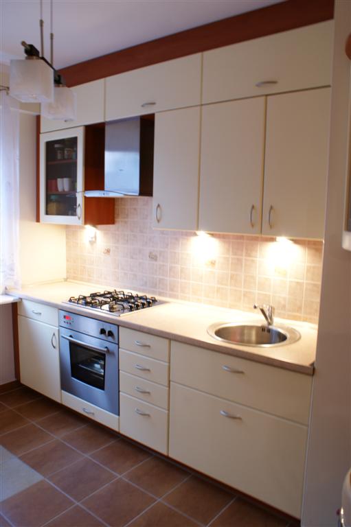 Jasna zabudowa kuchenna z blatem, okapem i szklaną szafką wiszącą, oświetlona punktowymi lampkami podszafkowymi i lampą sufitową, z brązową listwą pod sufitem i brązową terakotą na podłodze.